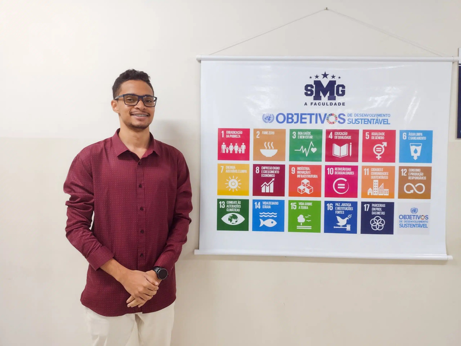 Thiago Felipe Moreira de Almeida, aluno de Engenharia da UNISMG, em foto ao lado do banner com os 17 ODS. O acadêmico desenvolveu uma telha ecológica para, inspirado pelo ODS 7. Foto: Sthefany Macedo/UNISMG.